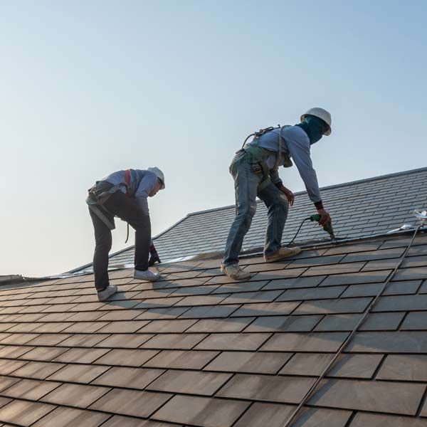 Two roofers wearing safety gear are working on a tiled roof on a sunny day.