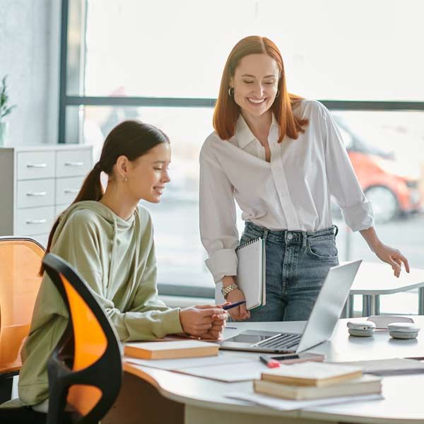 Two women in the office, one at a laptop, pointing, the other smiling, the tutor holding a notepad.