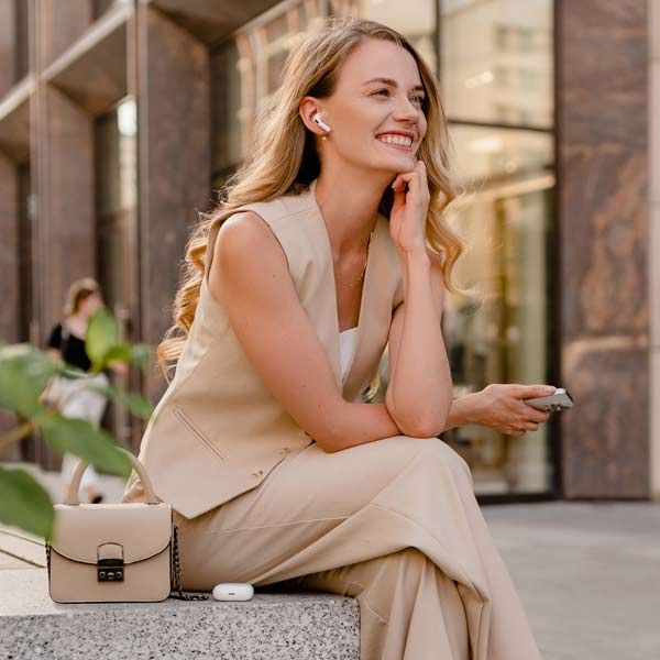 A professional model in a beige vest and trousers sits outdoors, wearing earbuds and holding a phone near a small handbag.
