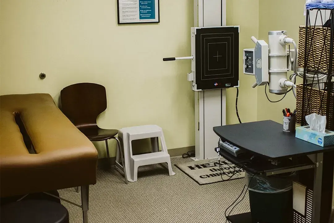 Medical exam room with tan exam table, brown chair, wall-mounted equipment, and black desk.