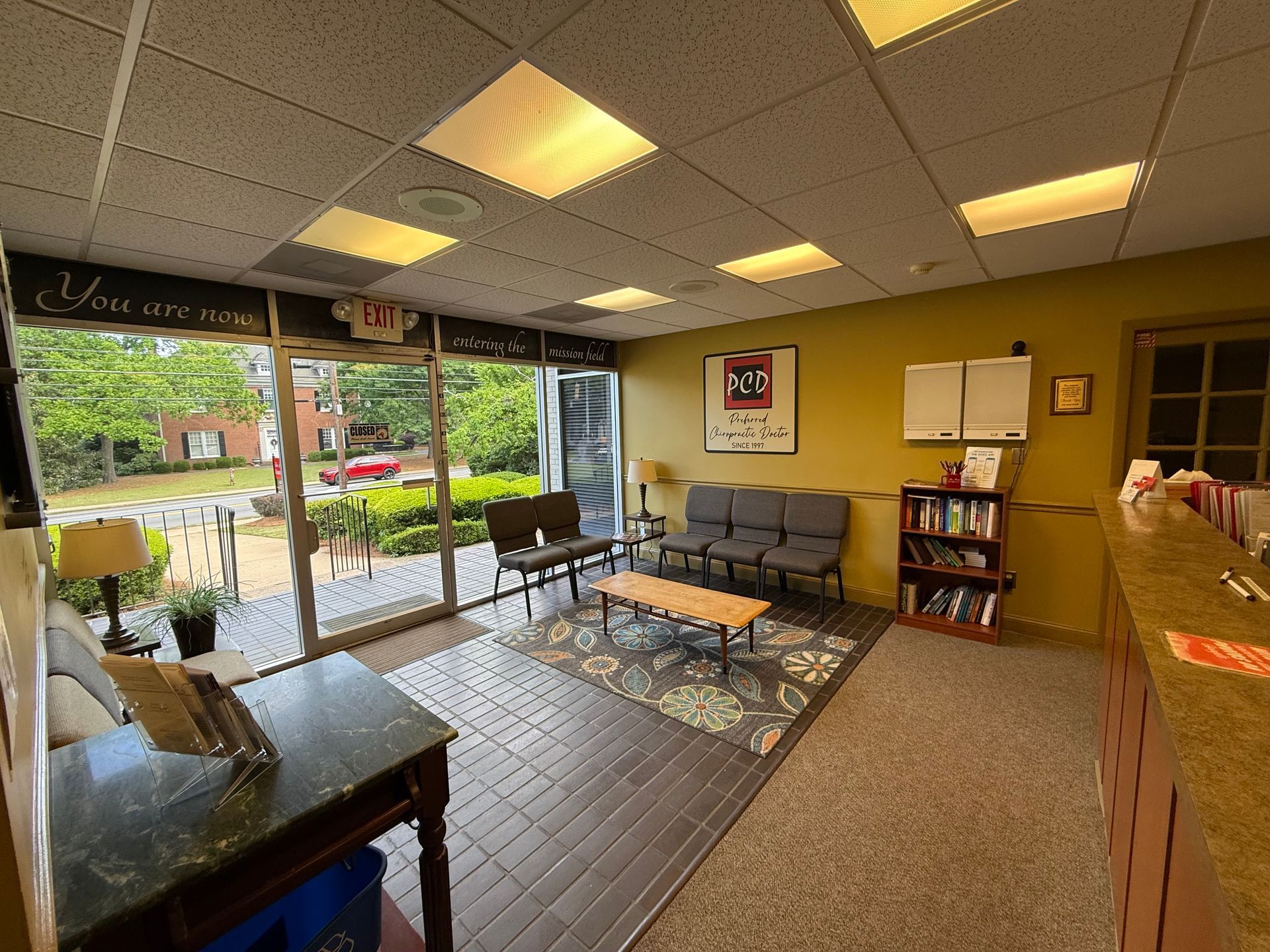 Lobby with tiled floor, couches, bookshelf, and large windows showing greenery outside.