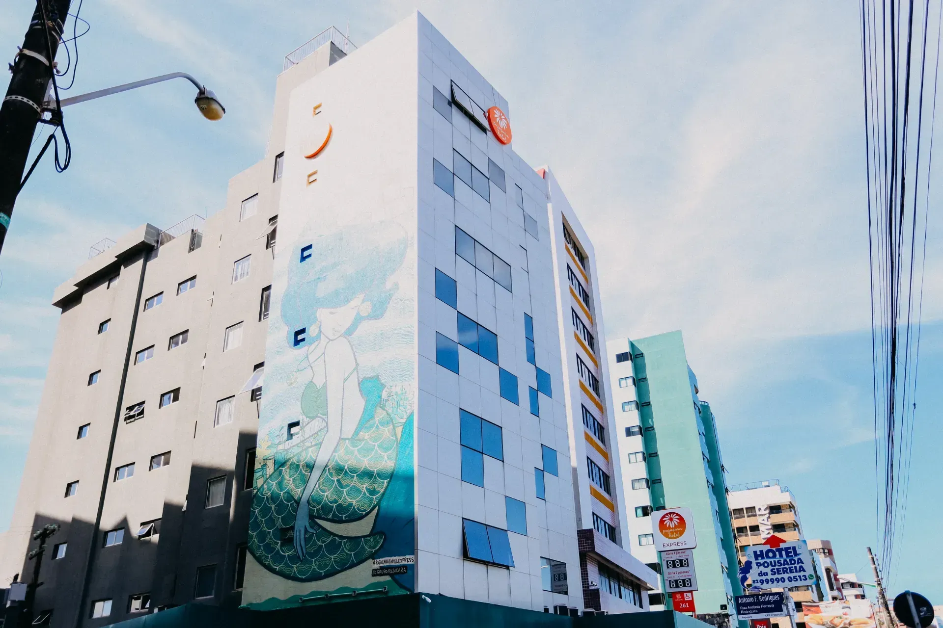 Prédios em um dia ensolarado. Prédio branco com janelas pixelizadas em azul e um mural. Outros prédios são visíveis.