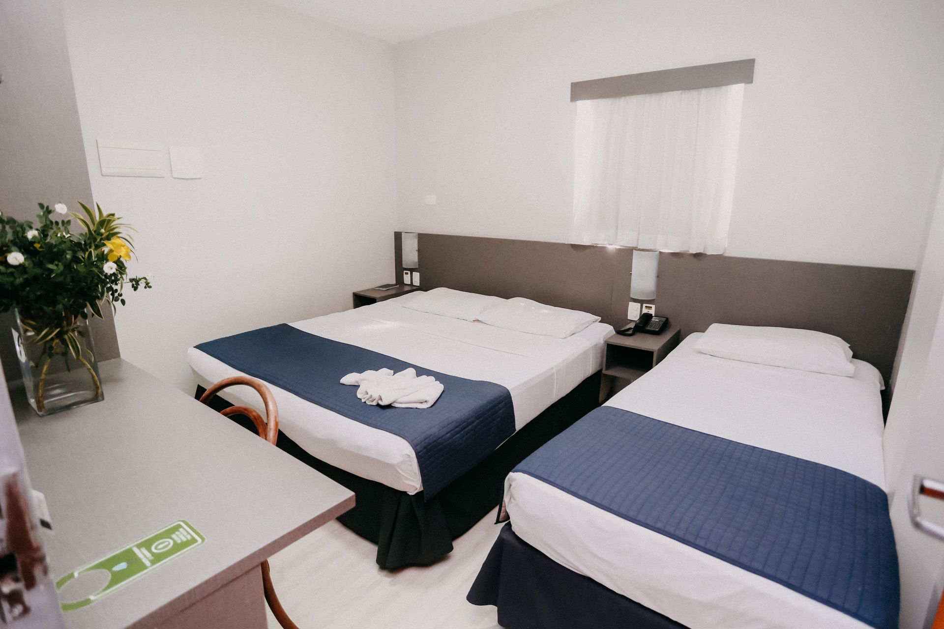 Quarto de hotel com duas camas, uma escrivaninha e uma janela. As camas têm peseiras azuis.