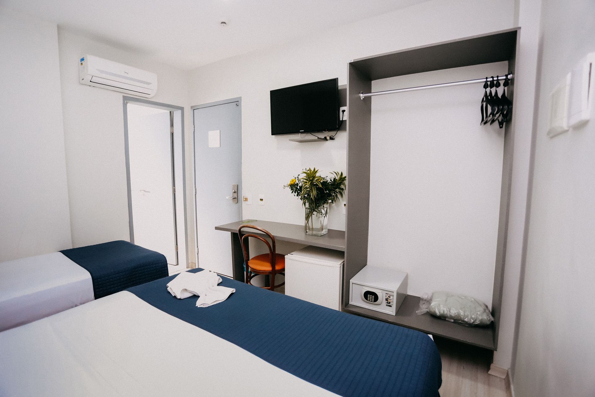Quarto de hotel com duas camas, escrivaninha, TV, guarda-roupa e frigobar.