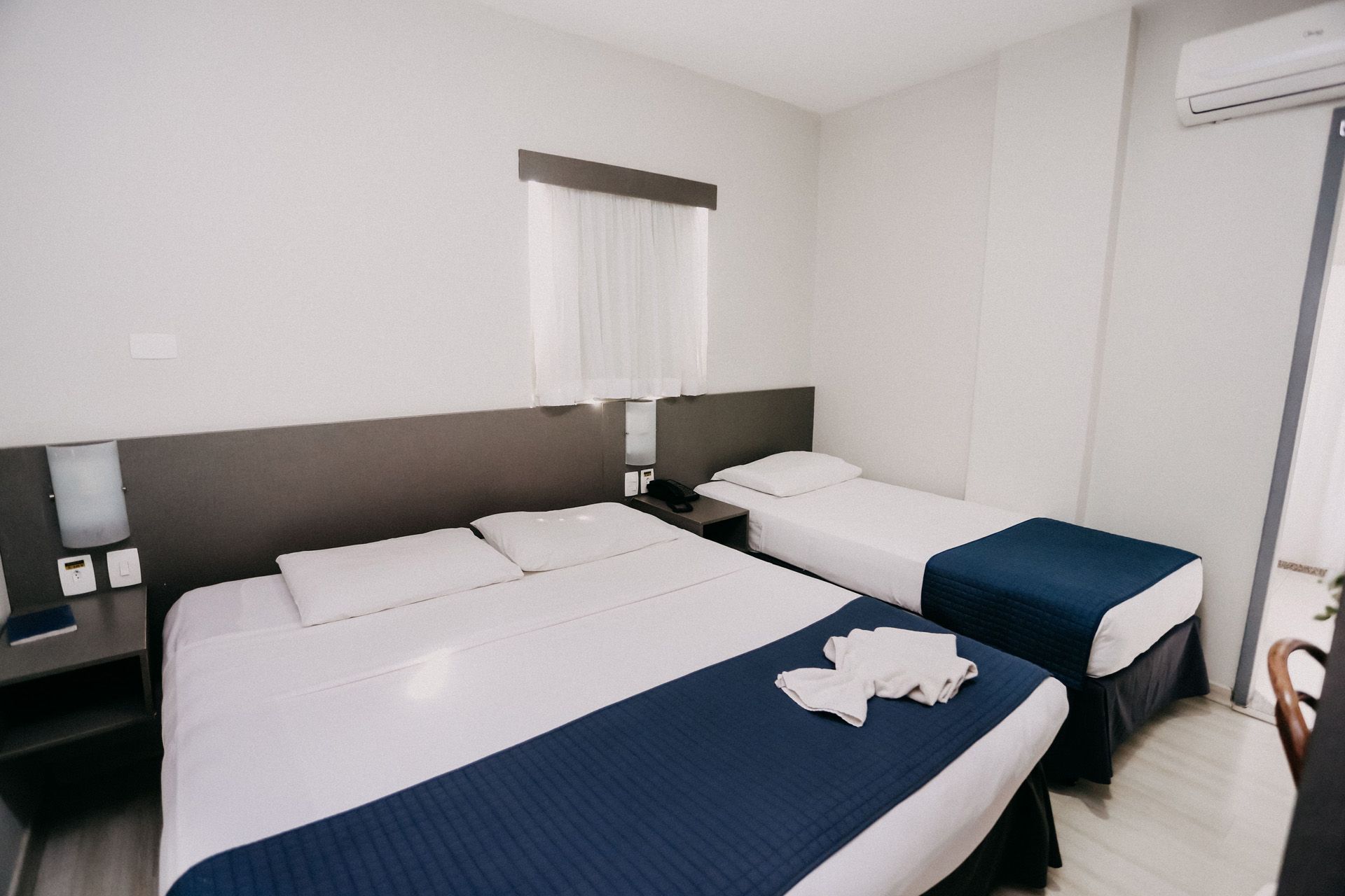 Quarto de hotel com uma cama de casal e uma cama de solteiro; paredes brancas, colchas azuis.