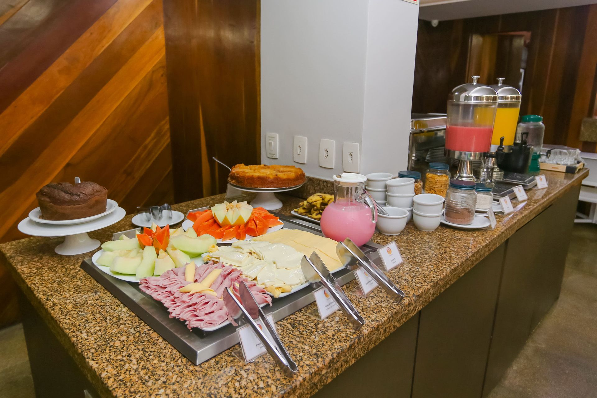 Buffet de café da manhã com diversos itens alimentares: pães, bolos, frutas e pratos quentes, em um refeitório.