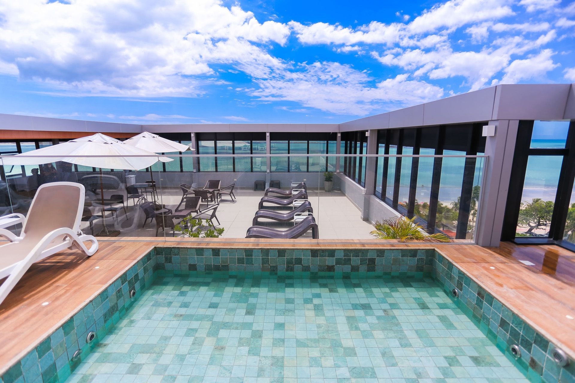 Piscina na cobertura com vista para o mar, espreguiçadeiras, mesas e guarda-sóis em um dia ensolarado com céu azul e algumas nuvens.