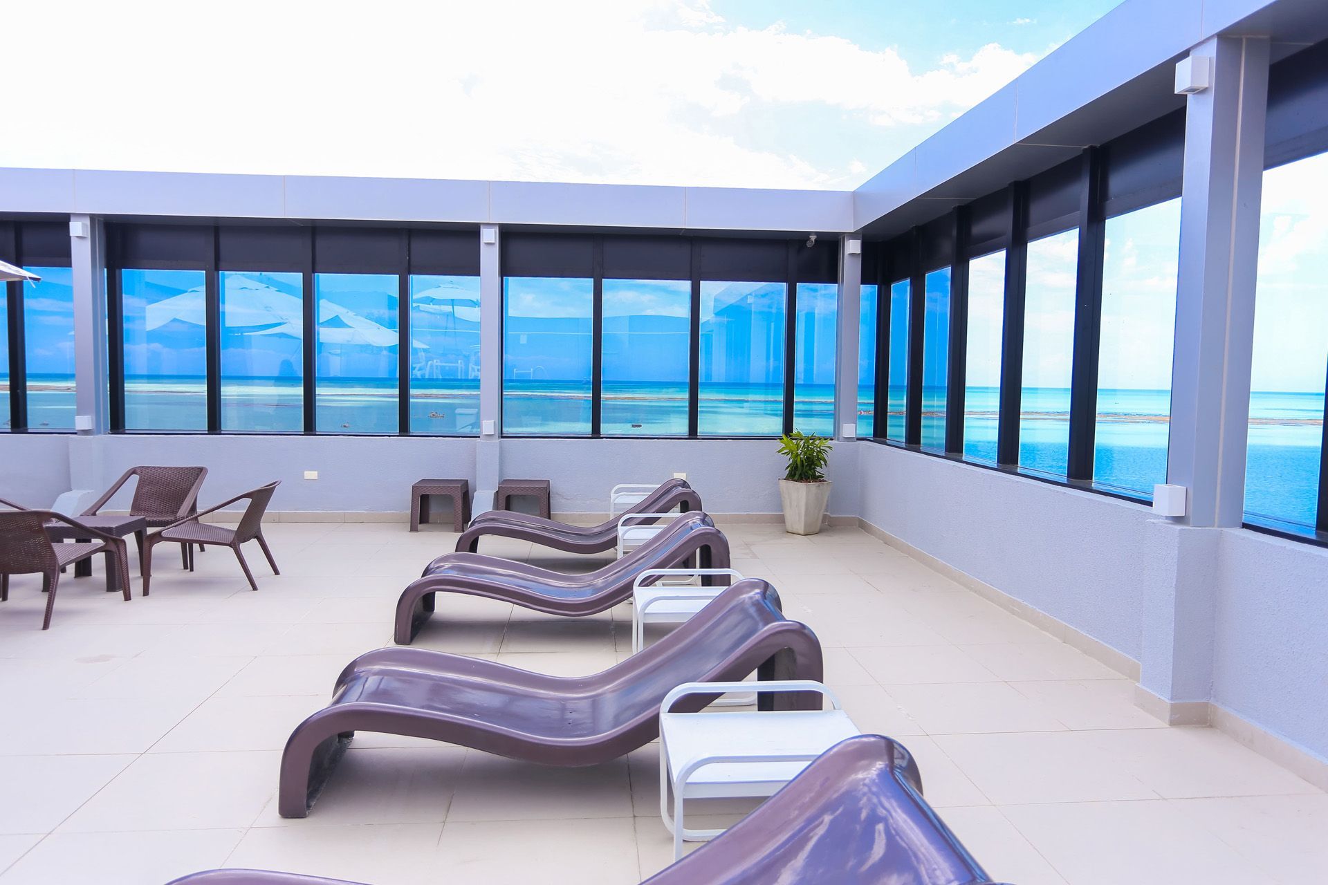 Salão na cobertura com espreguiçadeiras, mesas e vista para o mar através de grandes janelas com tonalidade azul.