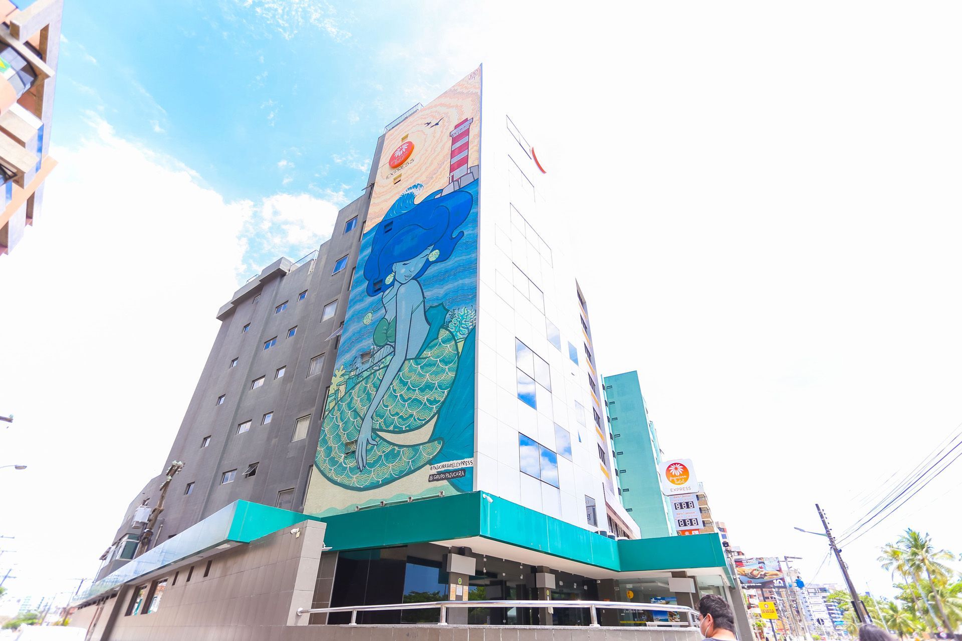 Prédio alto com mural de uma pessoa na água. Tons de turquesa e cinza com céu azul ao fundo.