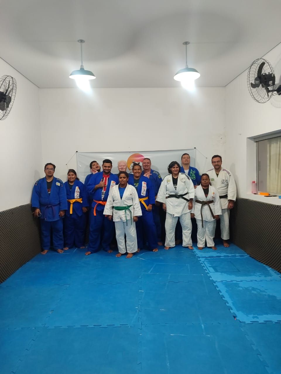Grupo de seis pesoas de pé uma do lado da outra no tatame, vestindo kimono e máscaras faciais.