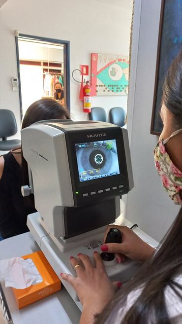 Médica oftamológica manipulando aparelho eletrônico que possue uma tela para melhor visualização do globo ocular, logo a sua frente  sentada está a paciente com o rosto sobre o aparelho.