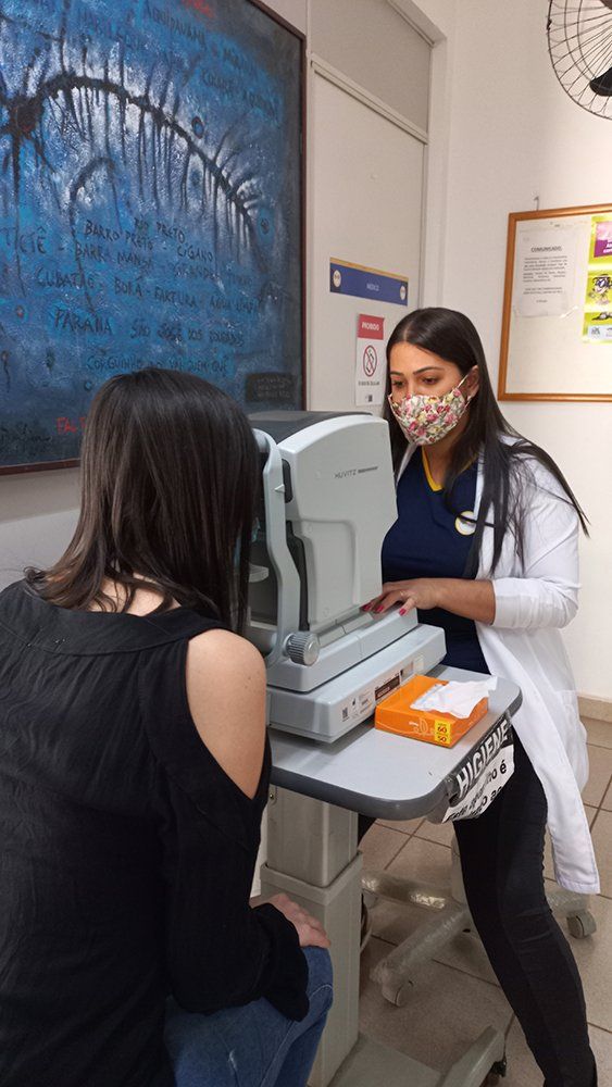 Médica oftalmológica manipulando equipamento clínico e examinando uma paciente que está sentada logo a sua frente.