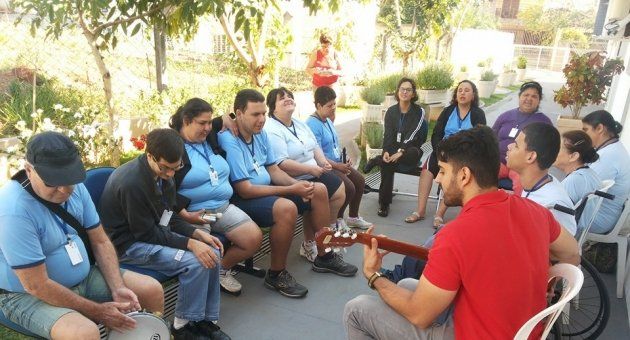 Um grupo de pessoas está sentado ao redor de um homem tocando violão.