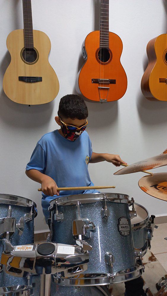 Menino usando óculos escuras e máscara facial tocando bateria com violões pendurados na parede ao fundo.