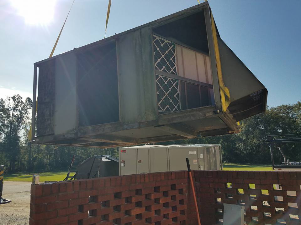 A large green HVAC unit is lifted by crane over a brick wall on a sunny day.