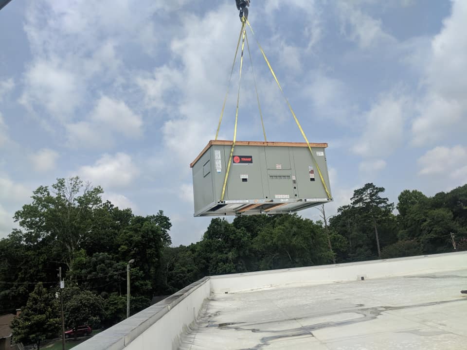 A large HVAC unit being lifted by a crane onto a rooftop. Overcast sky and trees in the background.