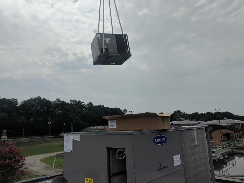 A crane lifts an air conditioning unit above another unit on a rooftop. Overcast sky, trees in the background.