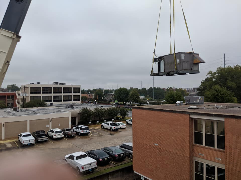 A crane lifting an HVAC unit onto a rooftop, with a parking lot and buildings in the background.