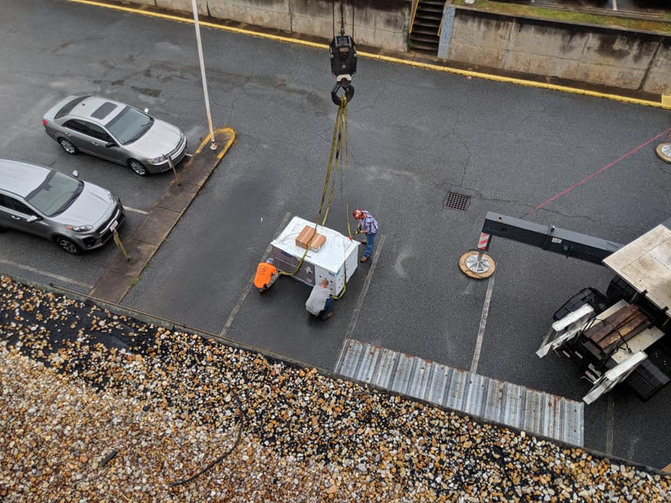 Crane lifting a covered box near parked cars and a worker on a parking lot.