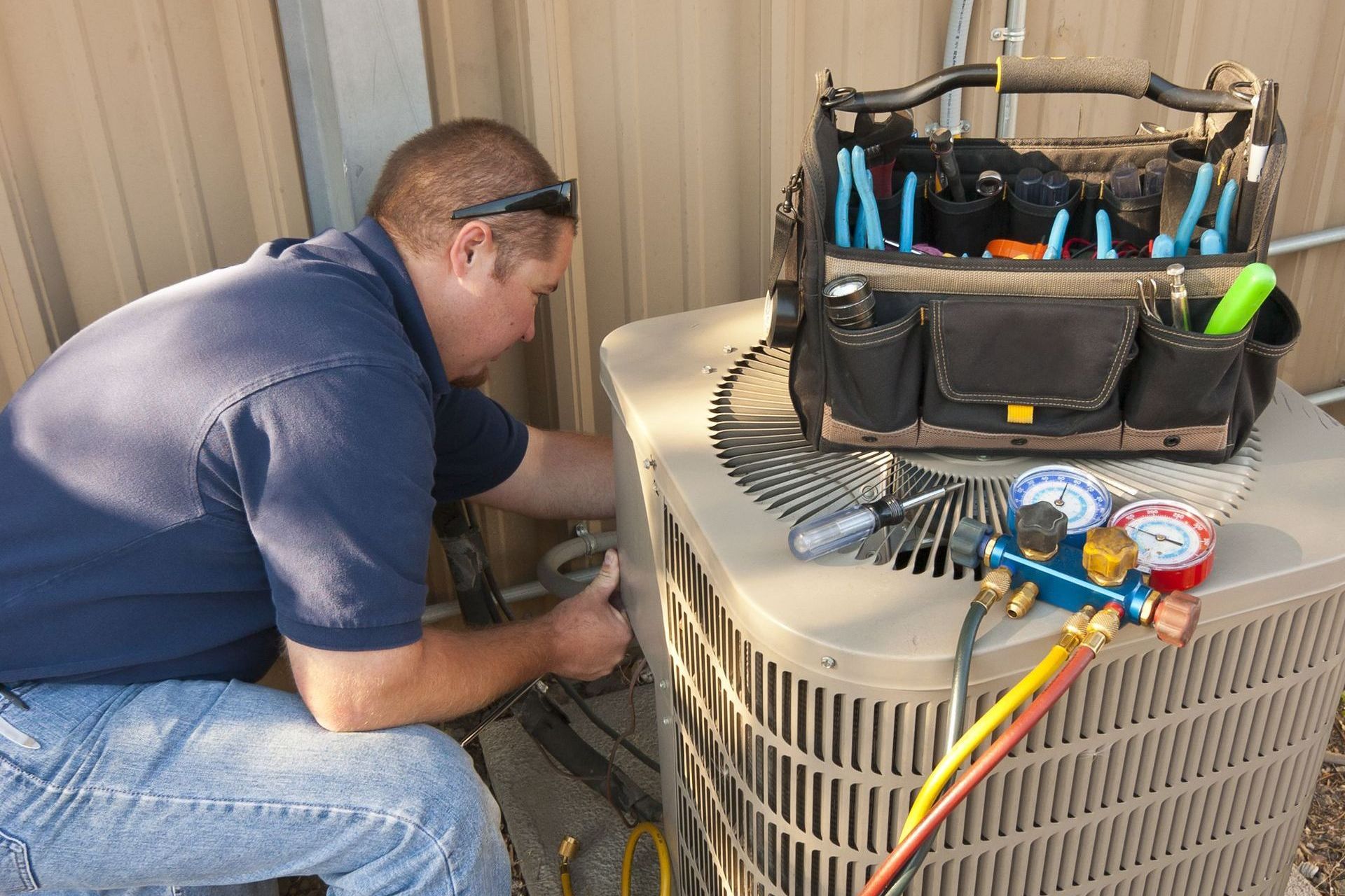HVAC technician kneels to repair an air conditioning unit outdoors, tool bag beside him.