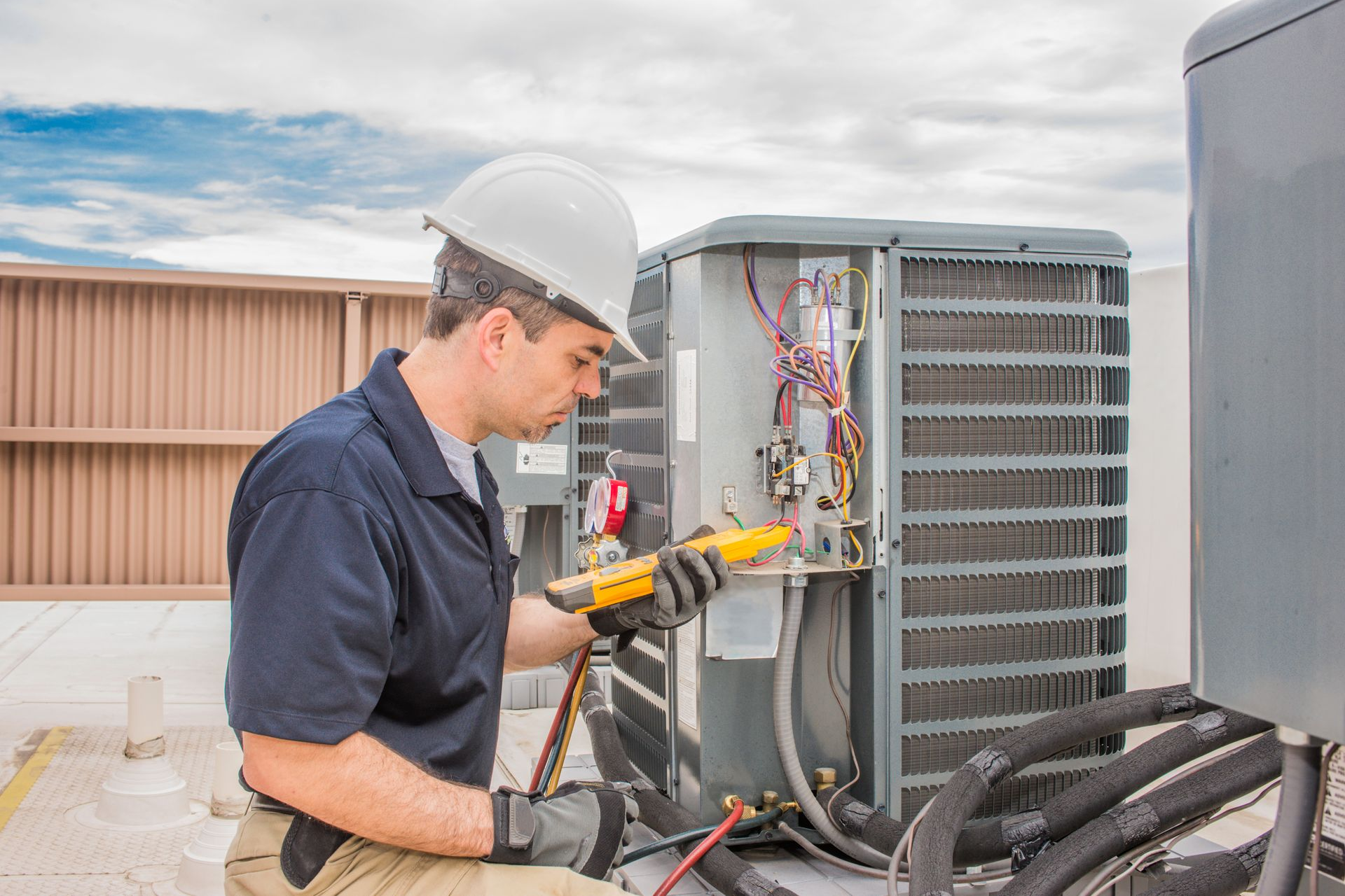 HVAC technician in a hard hat repairs an air conditioning unit outdoors, using a meter.