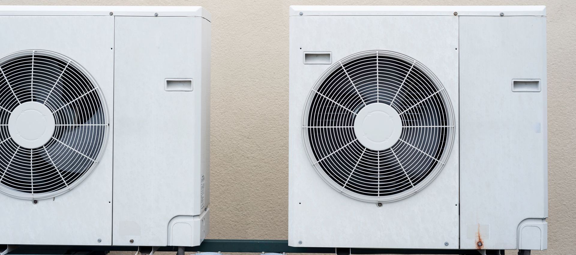 Two white air conditioning units mounted against a light beige wall.