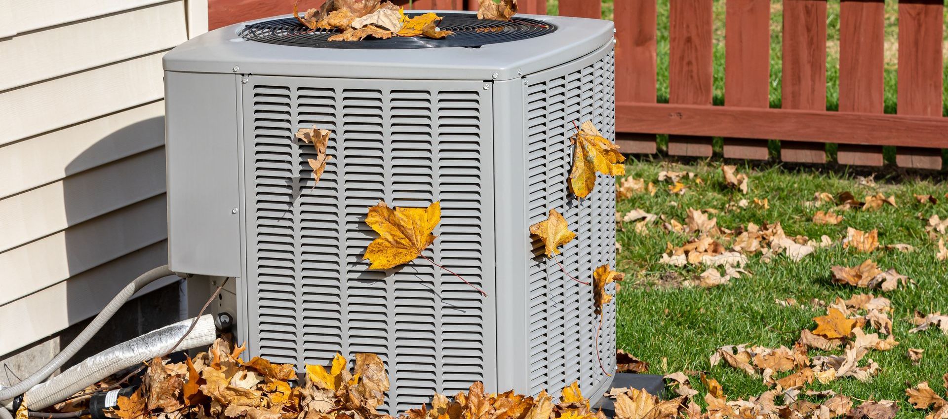 An air conditioning unit is covered in fallen leaves, located next to a house and a wooden fence.