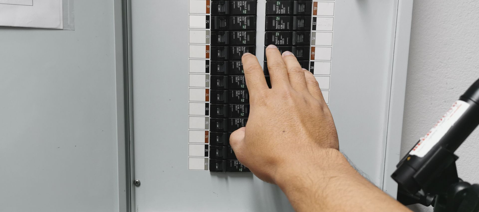 Person's hand reaching toward a circuit breaker panel. The panel is open, revealing multiple switches.