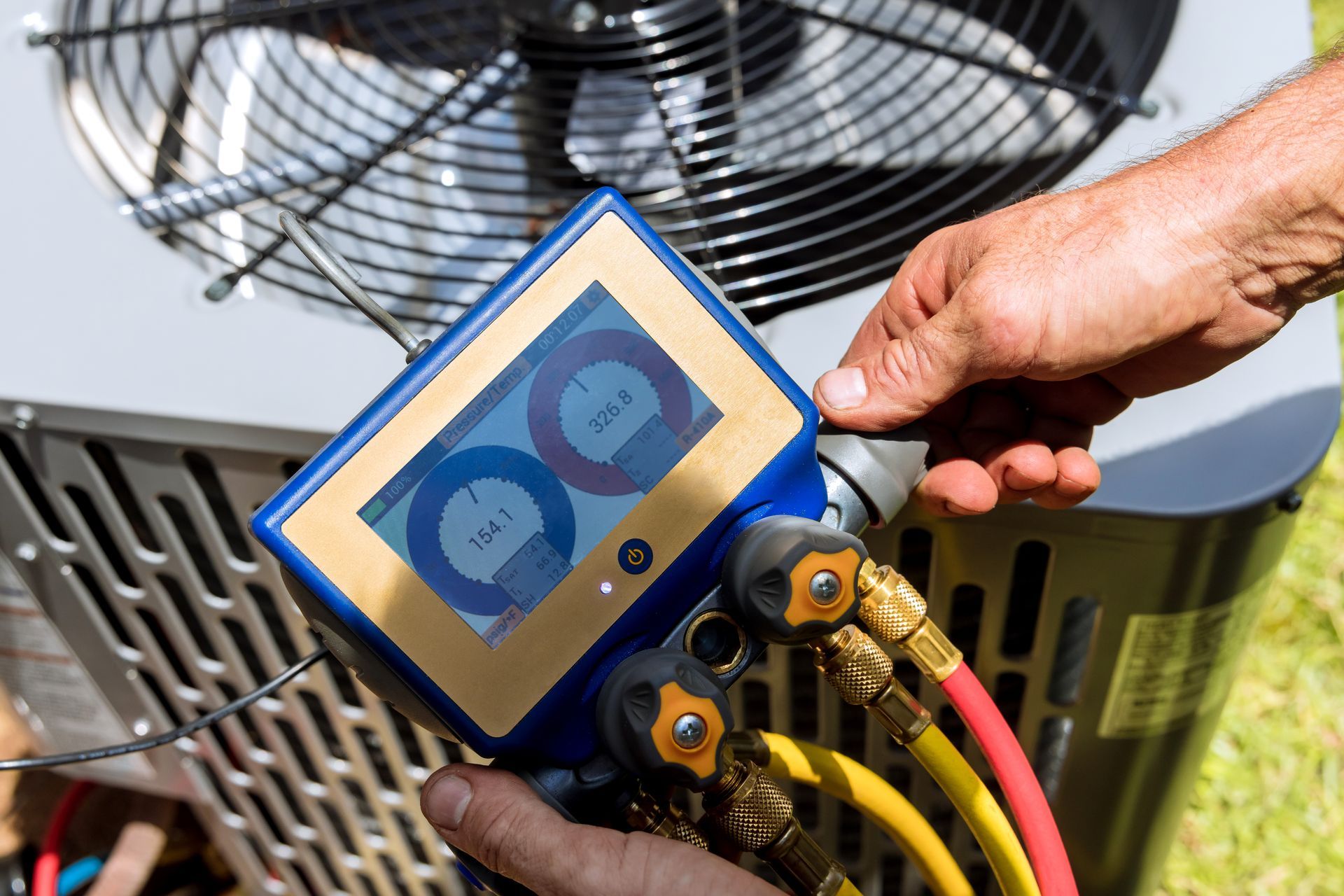 A technician checks AC pressure with a gauge. Blue, gold, red, and yellow hoses connect to the unit. Outdoors.