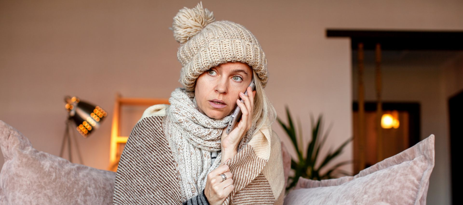 Person wrapped in blanket on couch, wearing hat and scarf, talking on phone with concerned expression.