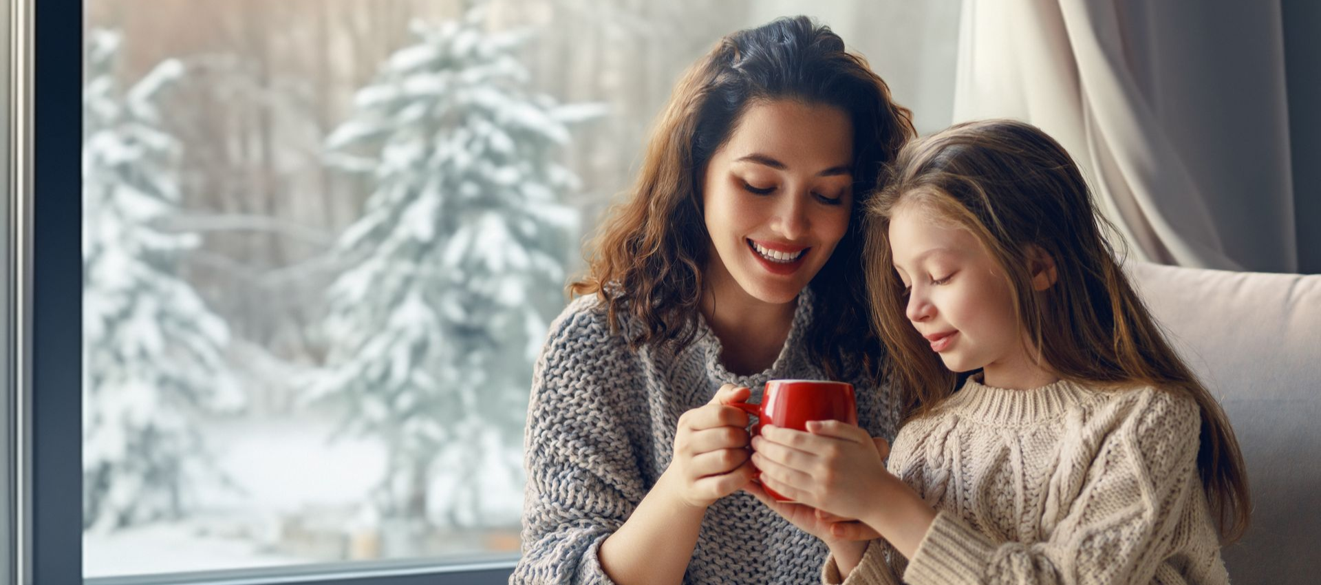 Woman and girl sharing a red mug, smiling, near a window with a snowy view.