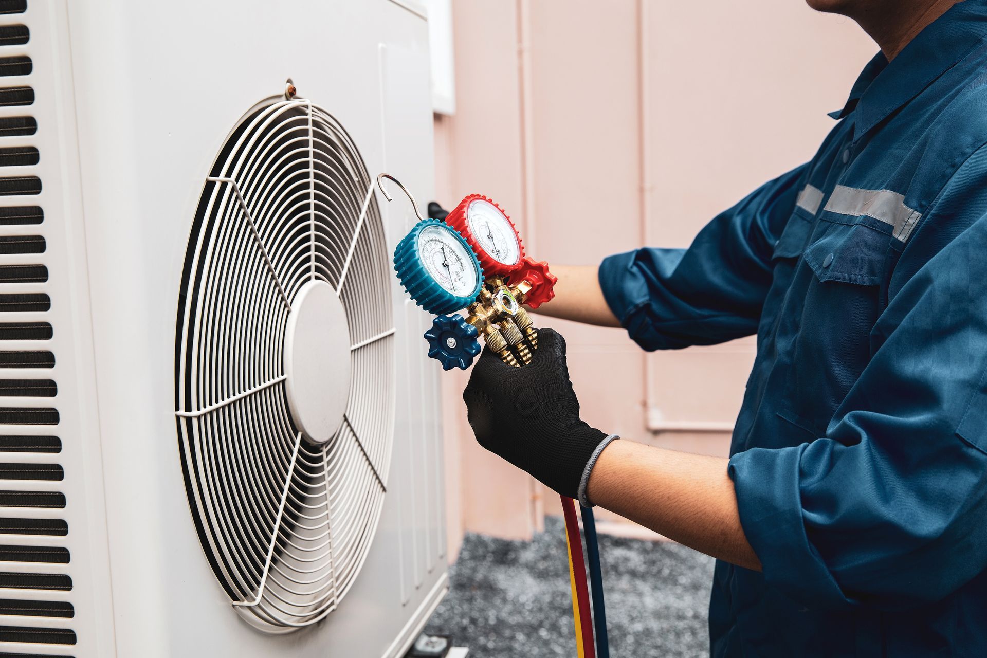 HVAC technician in blue jumpsuit uses gauges on an outdoor air conditioning unit with a large fan.