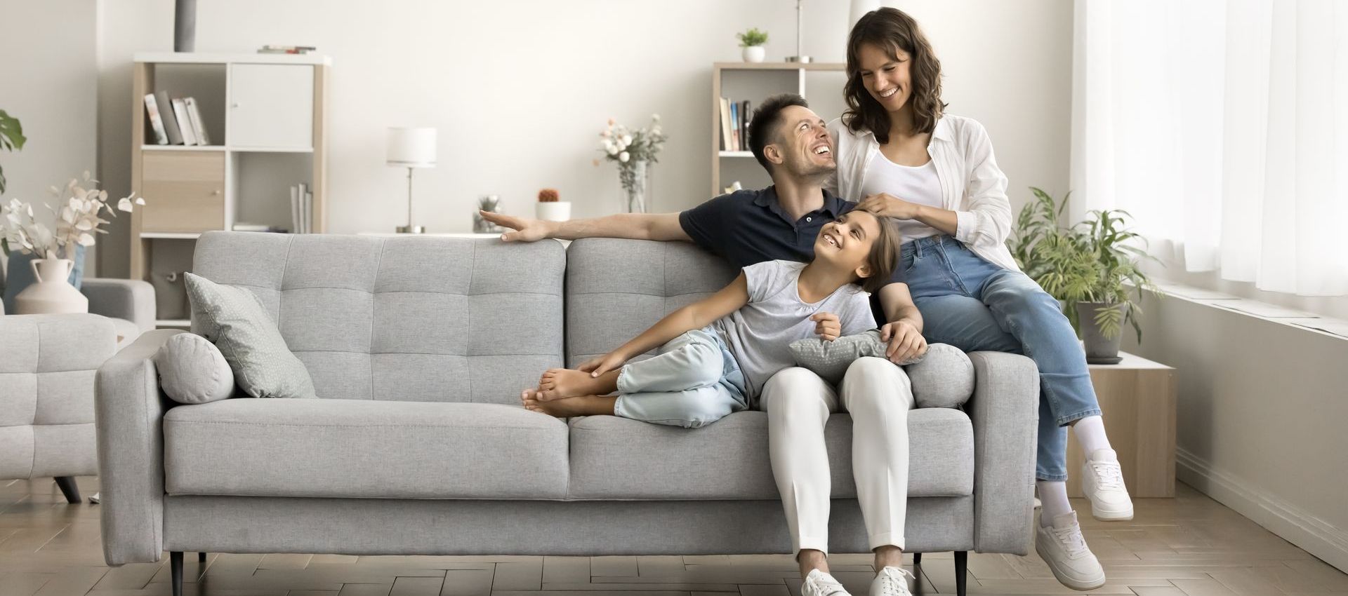A family of three is relaxing on a gray couch in a well-lit living room. The parents are smiling at their daughter.