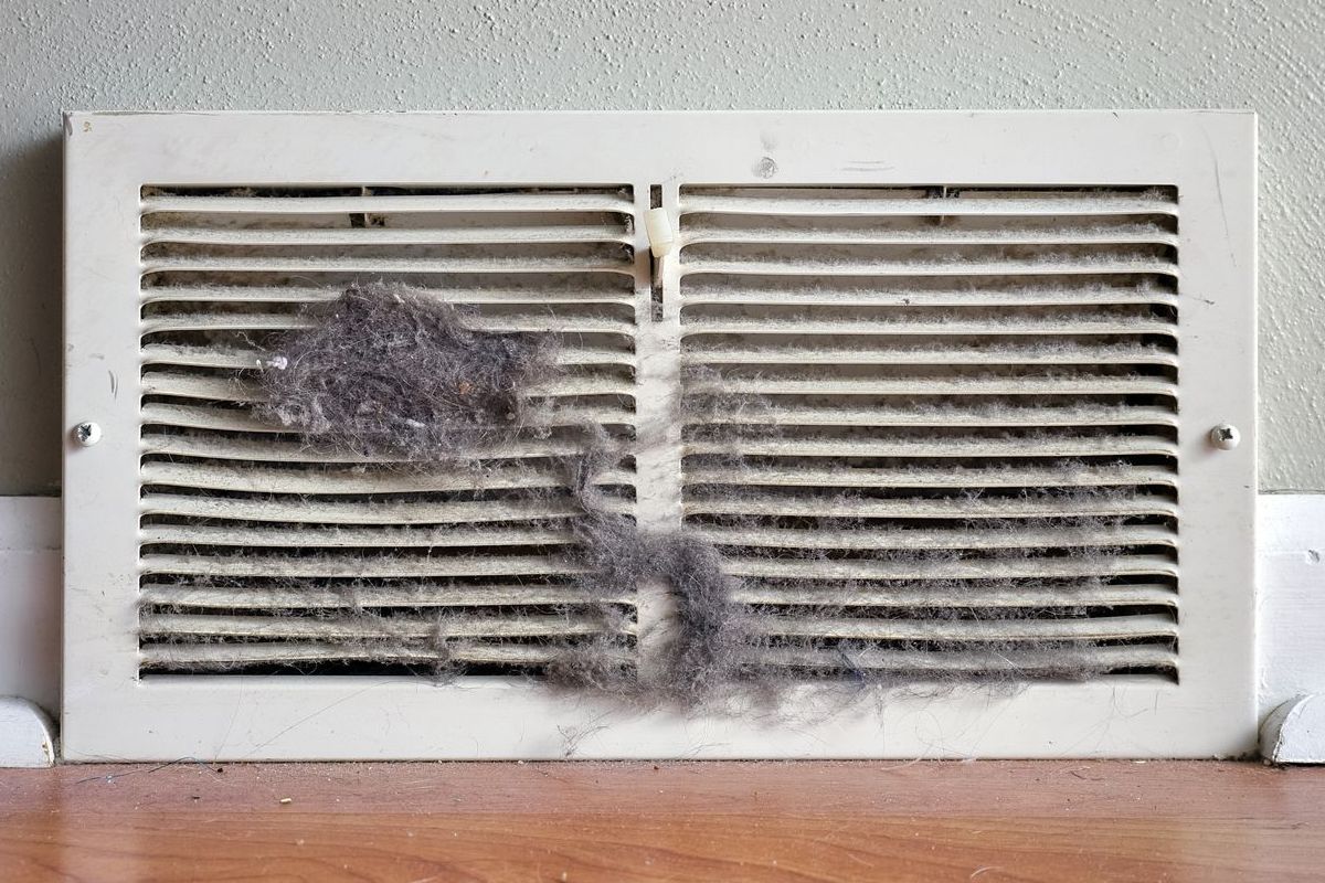 A dusty air vent on a wall above a wood floor; a large clump of dust partially blocks the vent.