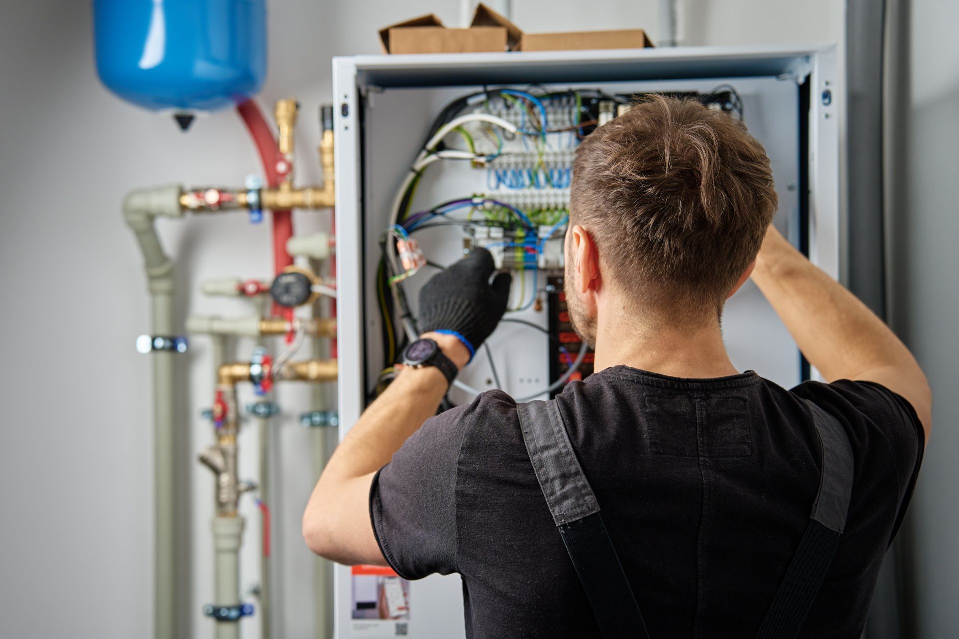 A man in black overalls repairs electrical wiring inside a white cabinet, plumbing visible on the left.