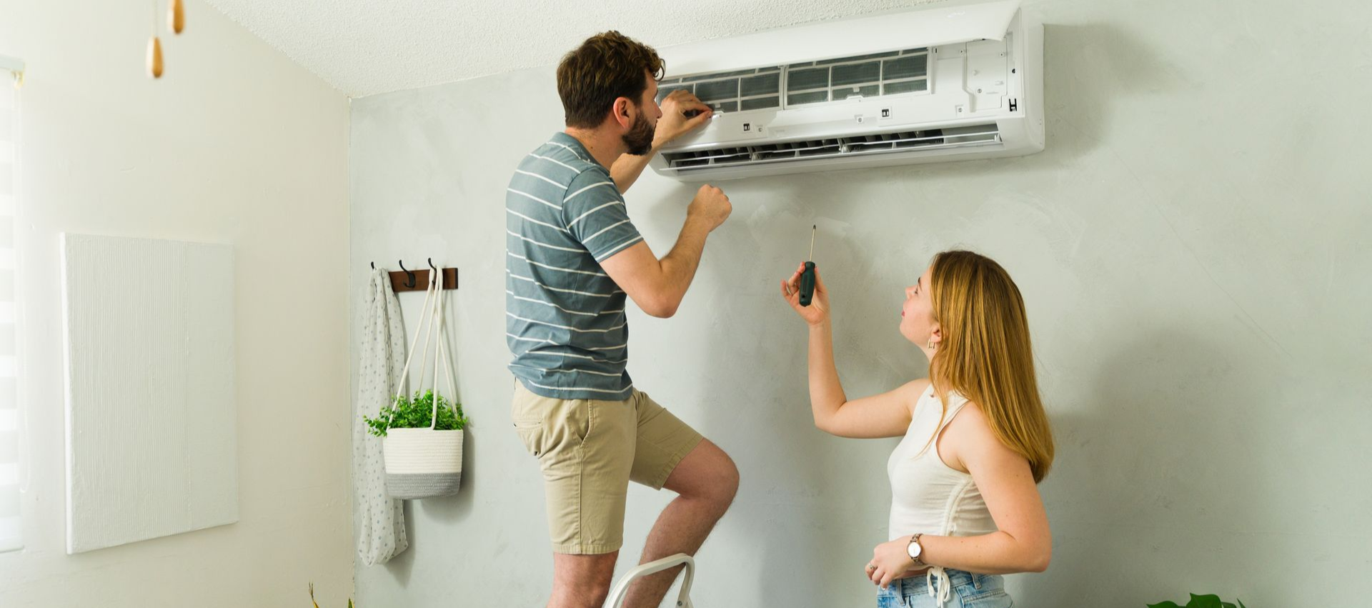 Man on ladder inspecting AC unit as woman holds remote, in a light-filled room.