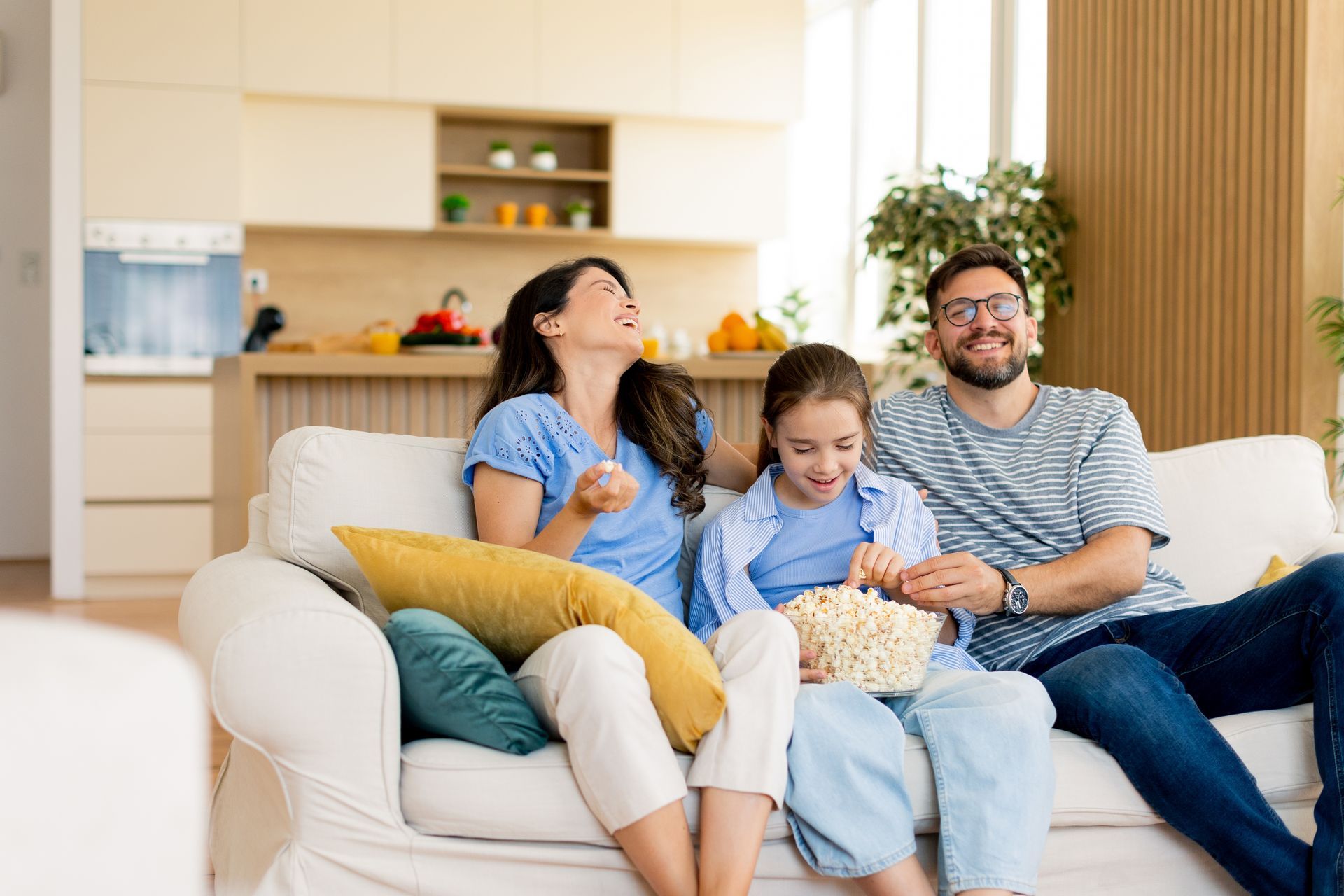 Family laughing while watching TV and eating popcorn on a white couch in a bright living room.