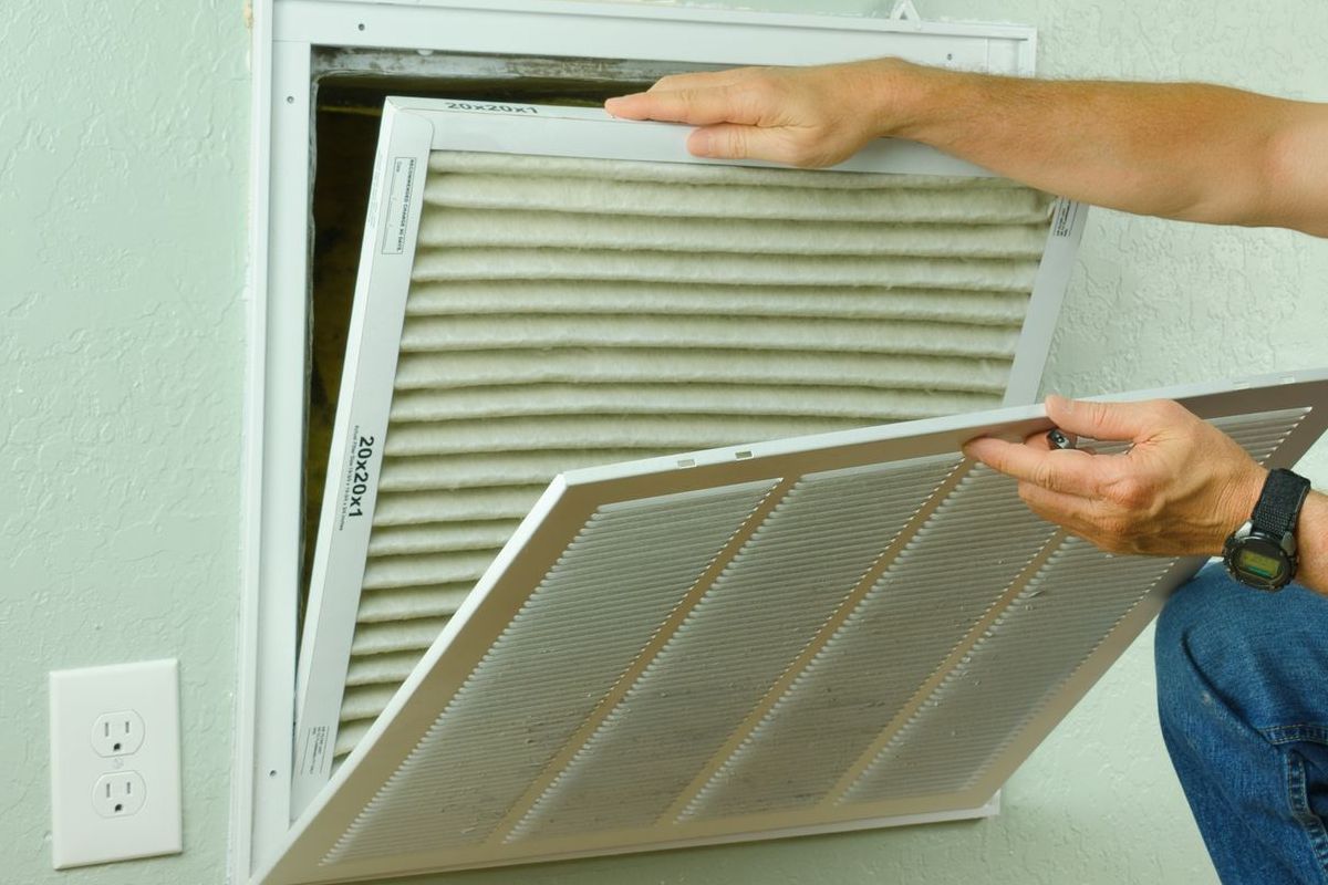 A man in a burgundy shirt is kneeling down to remove a filter from an air vent.