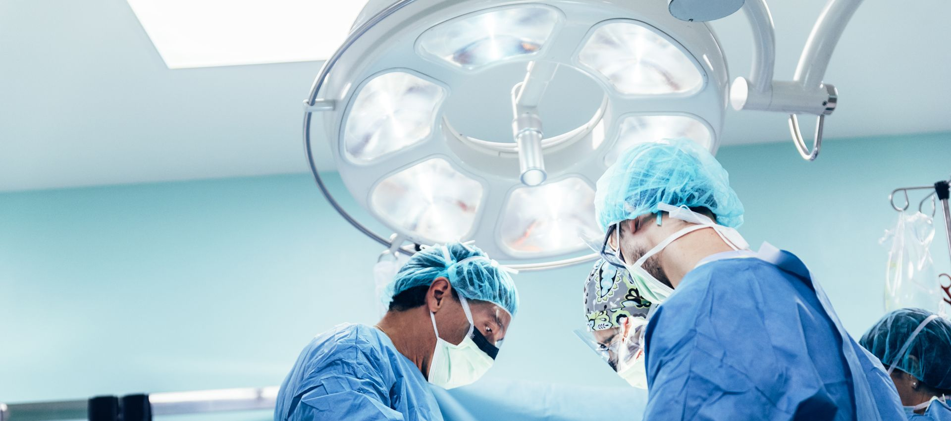 Surgeons in blue scrubs operate under bright lights in an operating room.