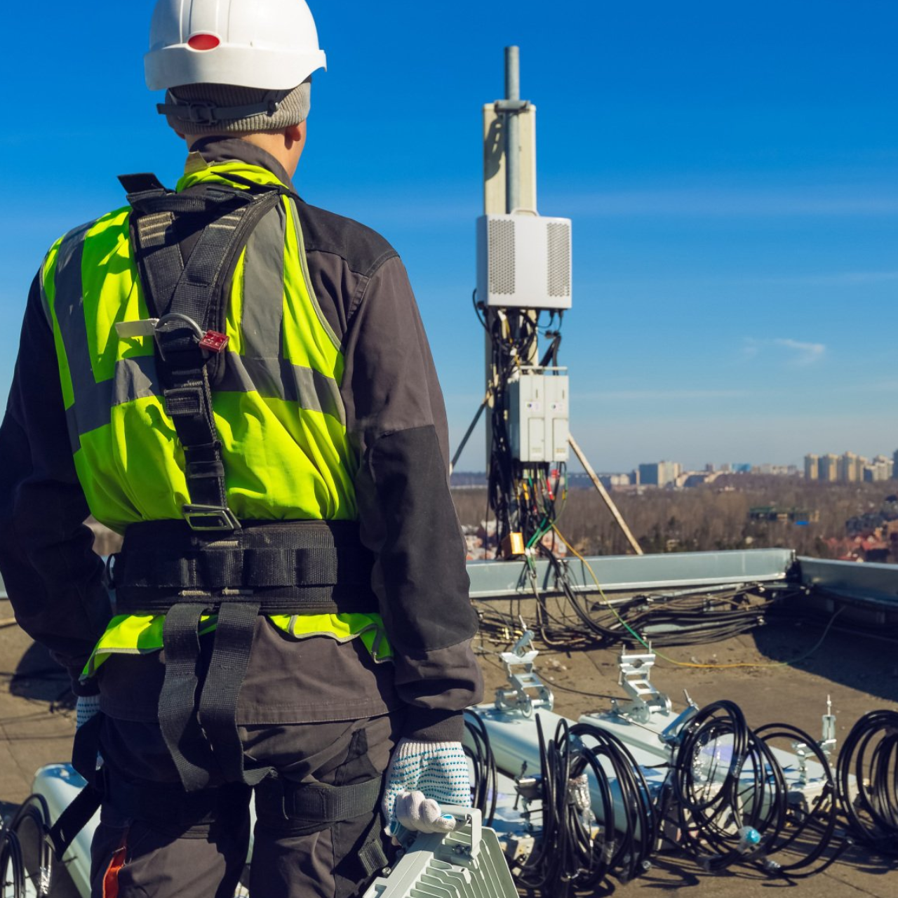 Worker in high vis jacket in foreground looking out over a city in the distance with antenna equipment layed out in front of him. 