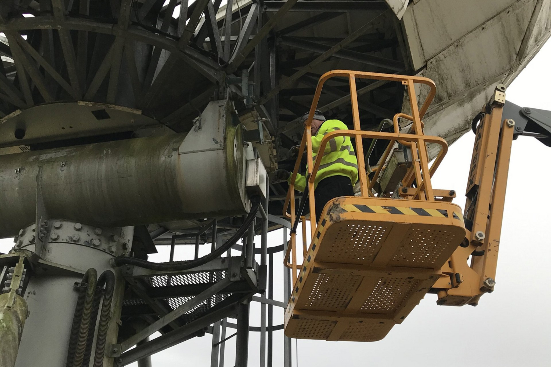 Working in high jacket in a cherry picker looking at the base of a satellite antenna dish,