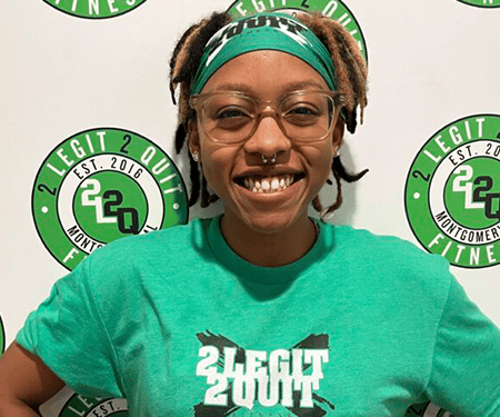 Smiling person in glasses and a green bandana, wearing a green “2Legit Youth” shirt, standing before a logo backdrop
