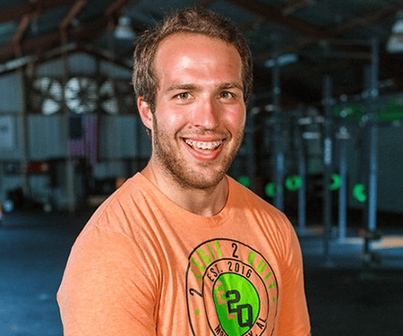 Smiling man in an orange T-shirt standing in a gym, with workout equipment in the background