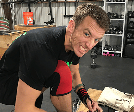 Man in black shirt and red shorts tying shoelaces in a gym, leaning forward and smiling.