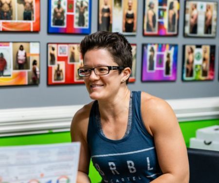 Smiling person with glasses in a blue tank top, seated indoors before a wall of framed photos