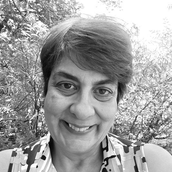 Smiling woman with short hair, patterned top, against leafy background.
