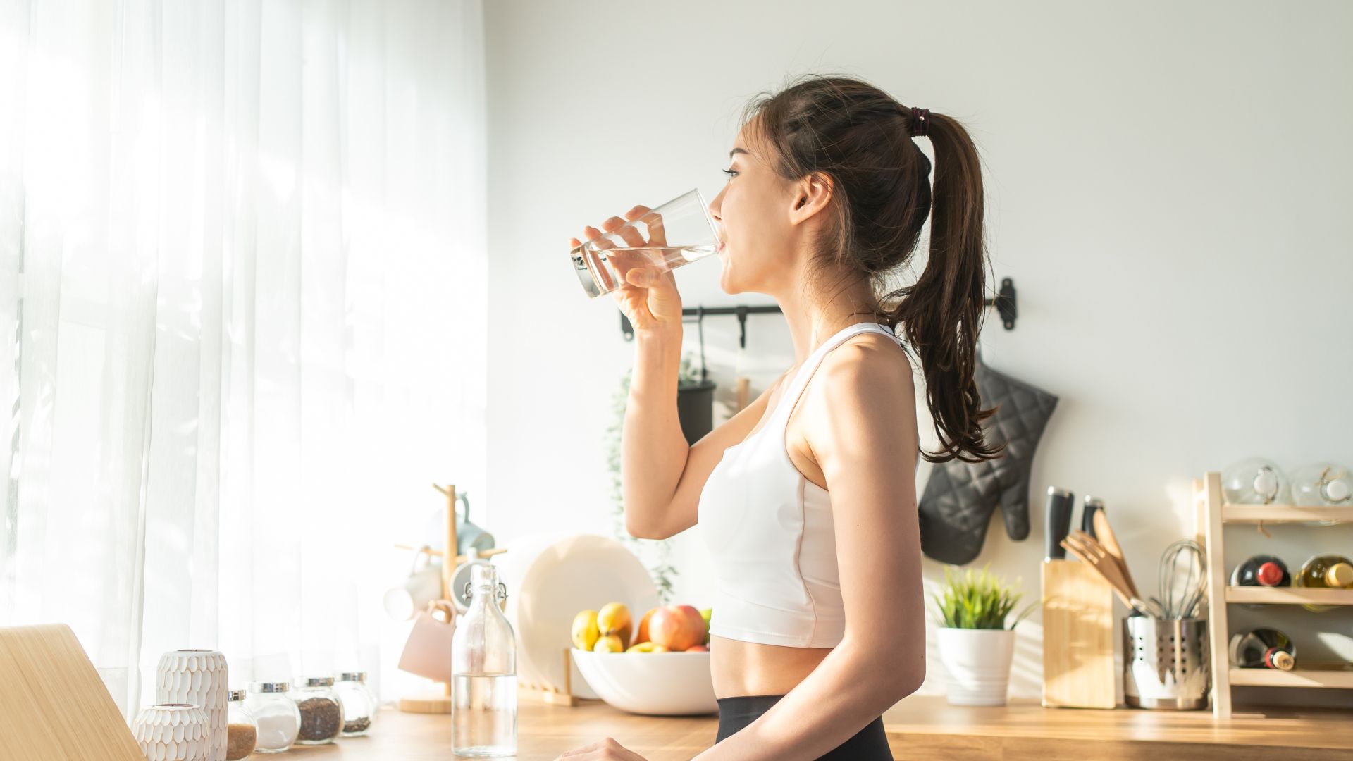 A woman is drinking water from a glass in a kitchen.