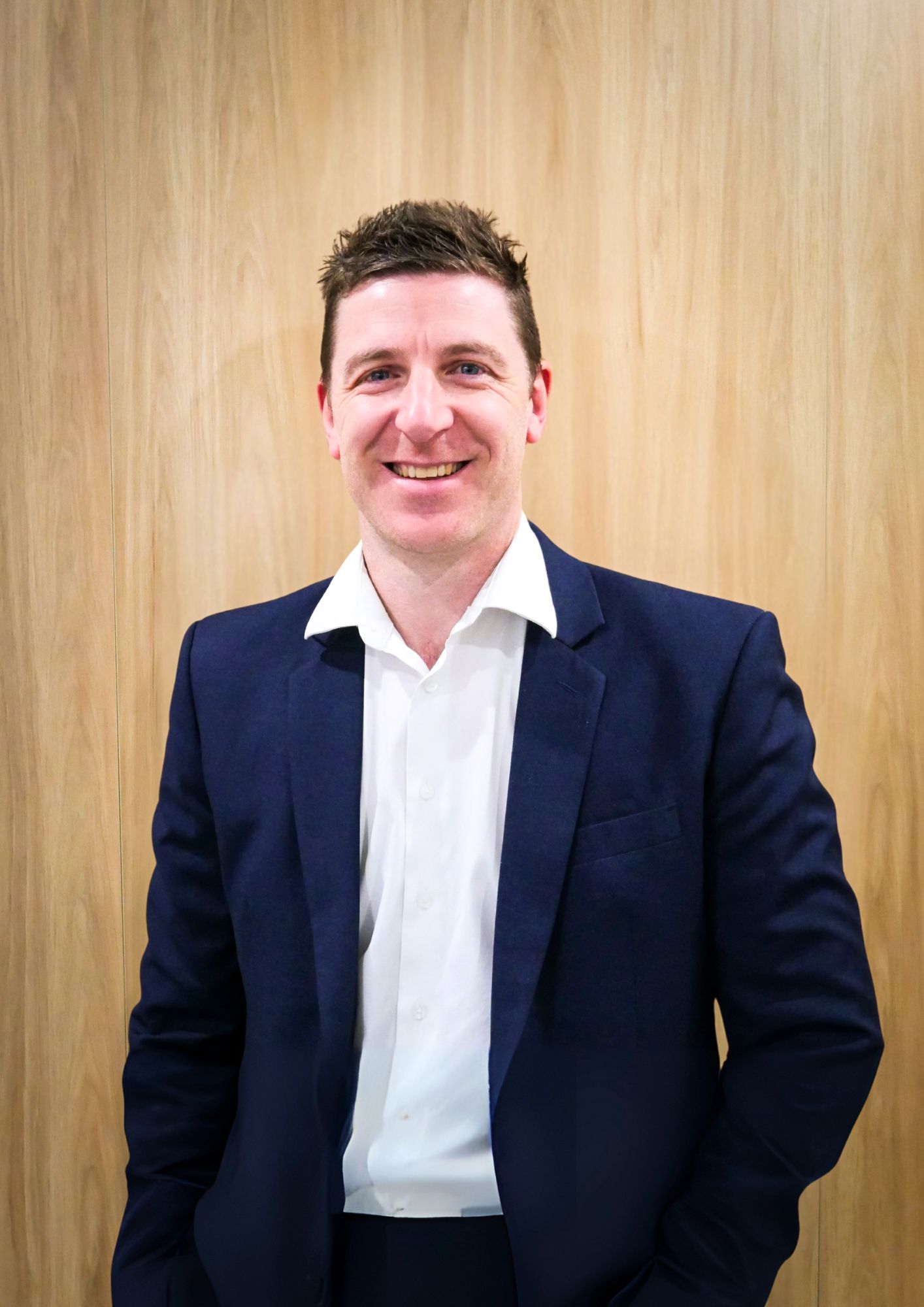 A smiling person wearing a white collared shirt and dark blue blazer, positioned against a light wood-paneled wall.