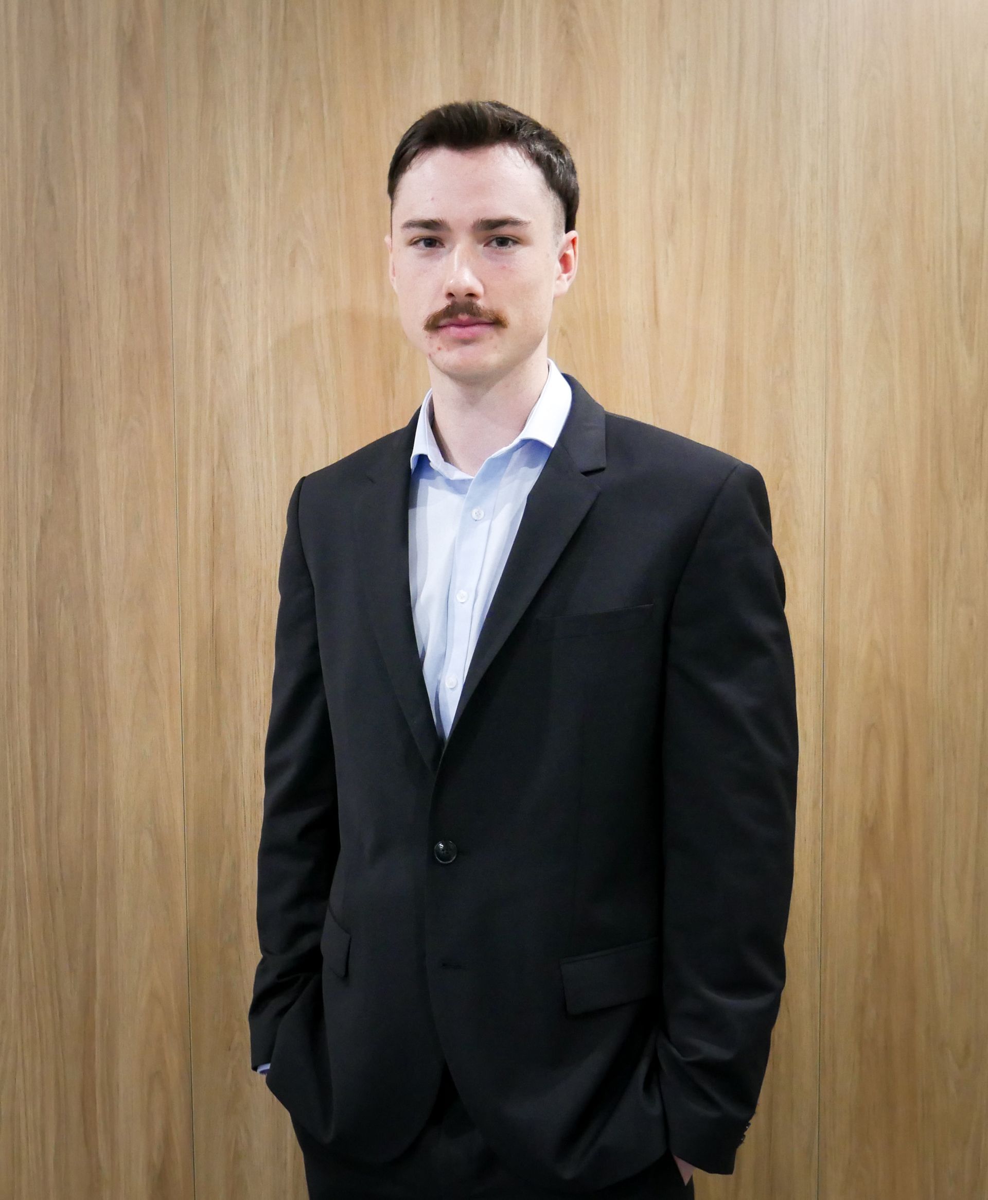 A person in a black suit and light blue collared shirt stands with hands in pockets against a wood-paneled wall.