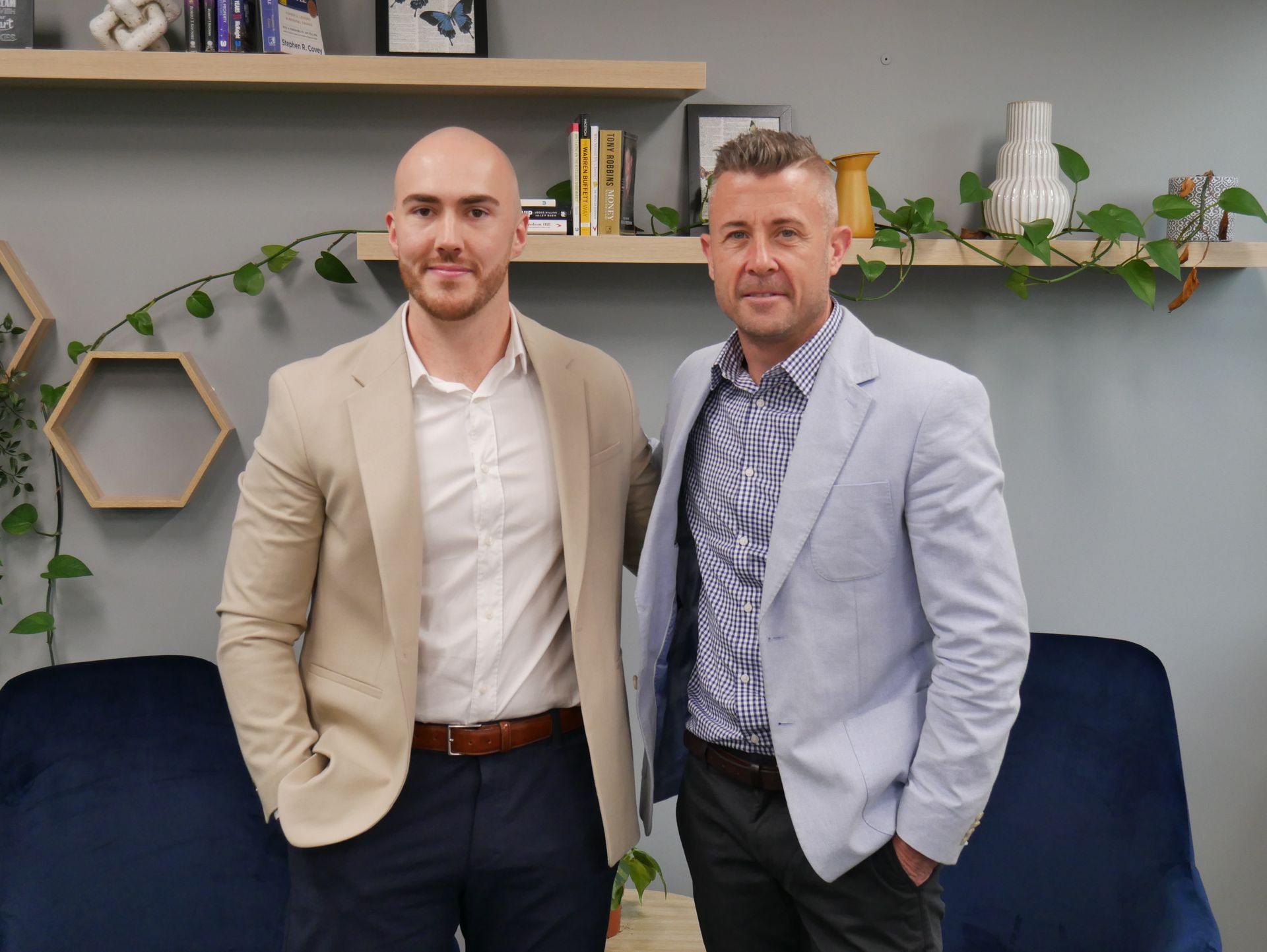 Two men in blazers standing side-by-side in an office setting with wall-mounted shelves, plants, and blue chairs.